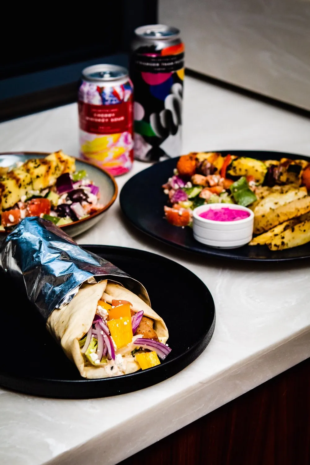 Wrapped gyro sandwich with grilled chicken, vegetables, and onions on a black plate, with two plates of Mediterranean salad and grilled potato wedges, and two colorful soda cans in the background.