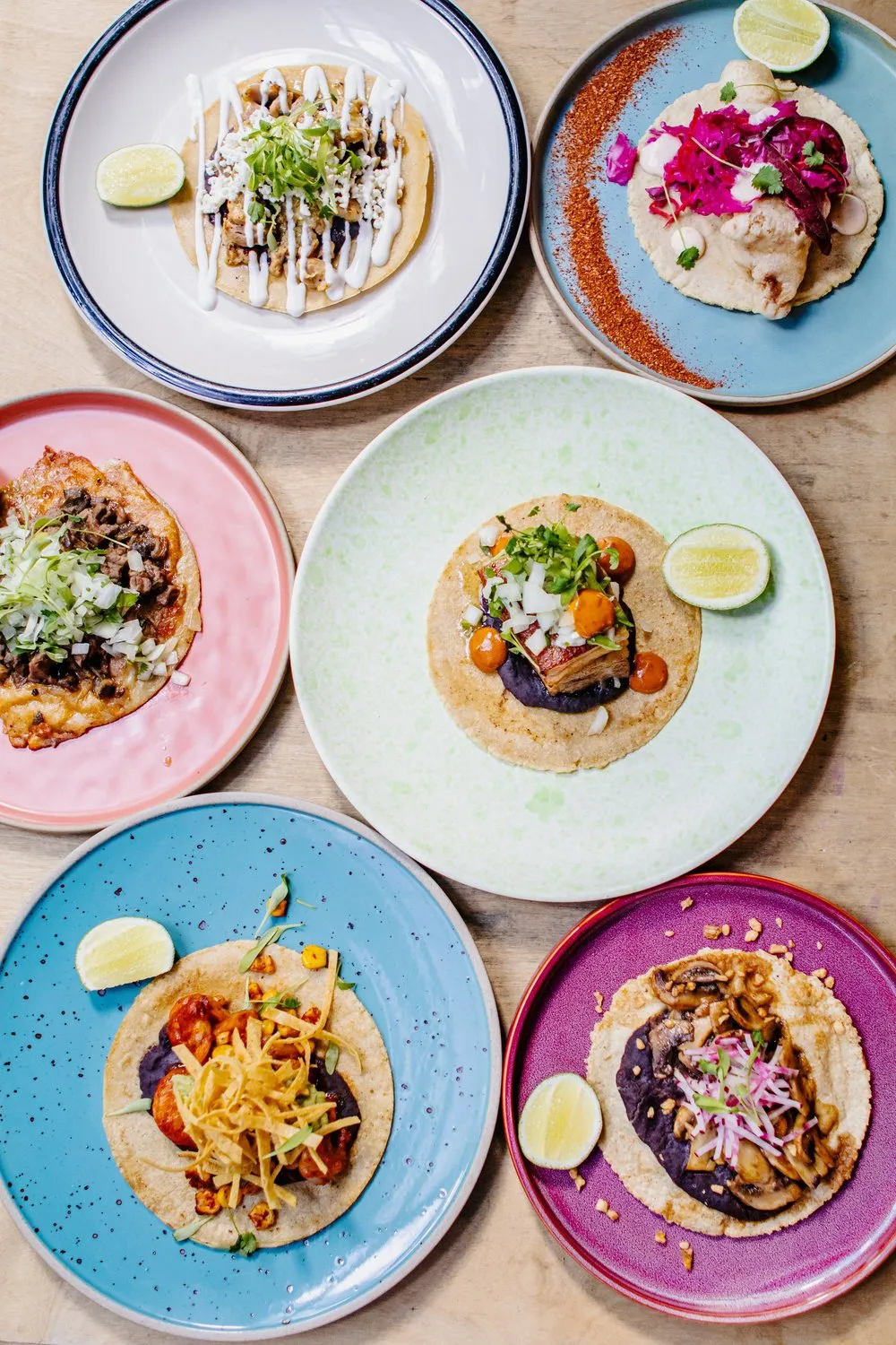 Six colorful plates, each with a different gourmet taco topped with various ingredients and garnished with lime wedges on a wooden table.