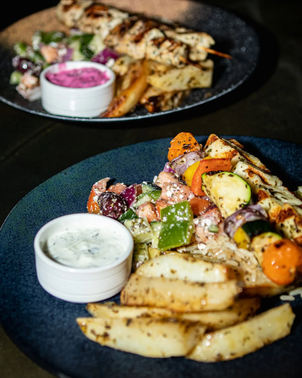Close-up of grilled vegetable skewers, seasoned potato wedges, Greek salad, and creamy sauces on blue and black plates.
