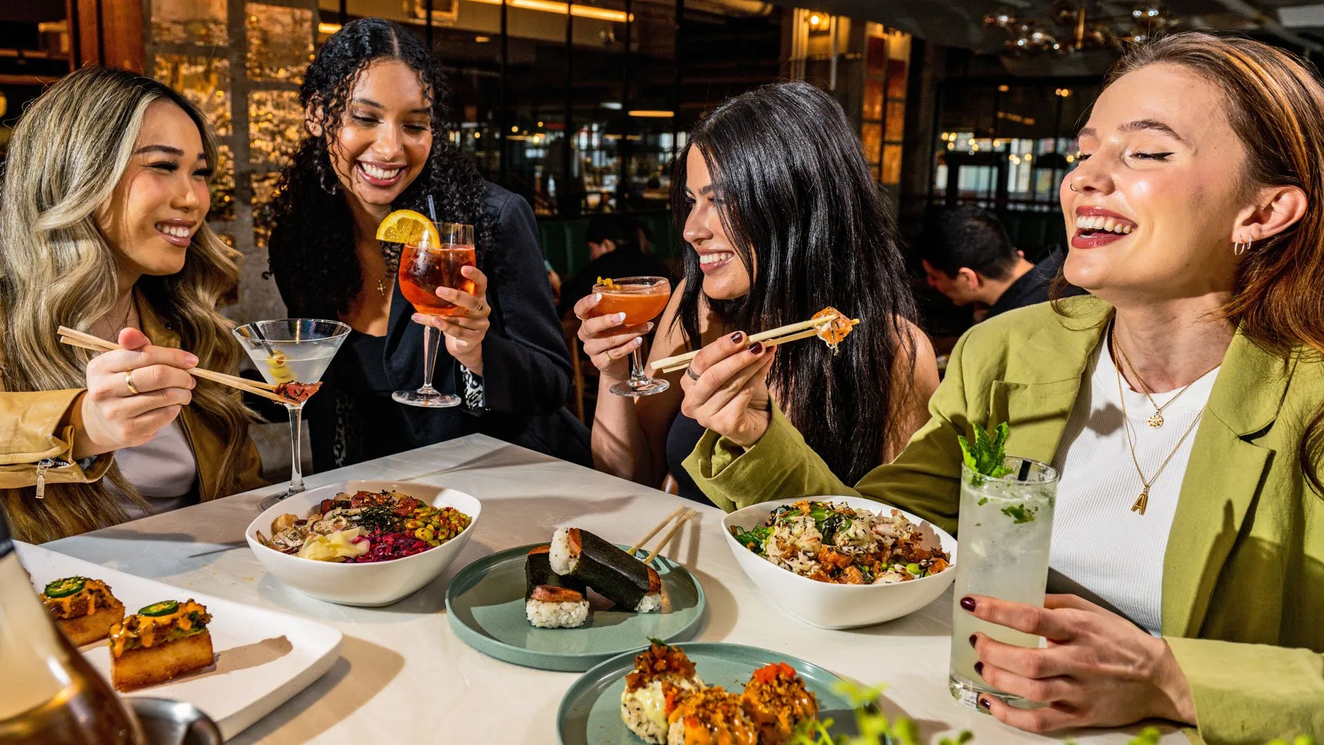 Four women enjoying sushi and cocktails together at a restaurant table.