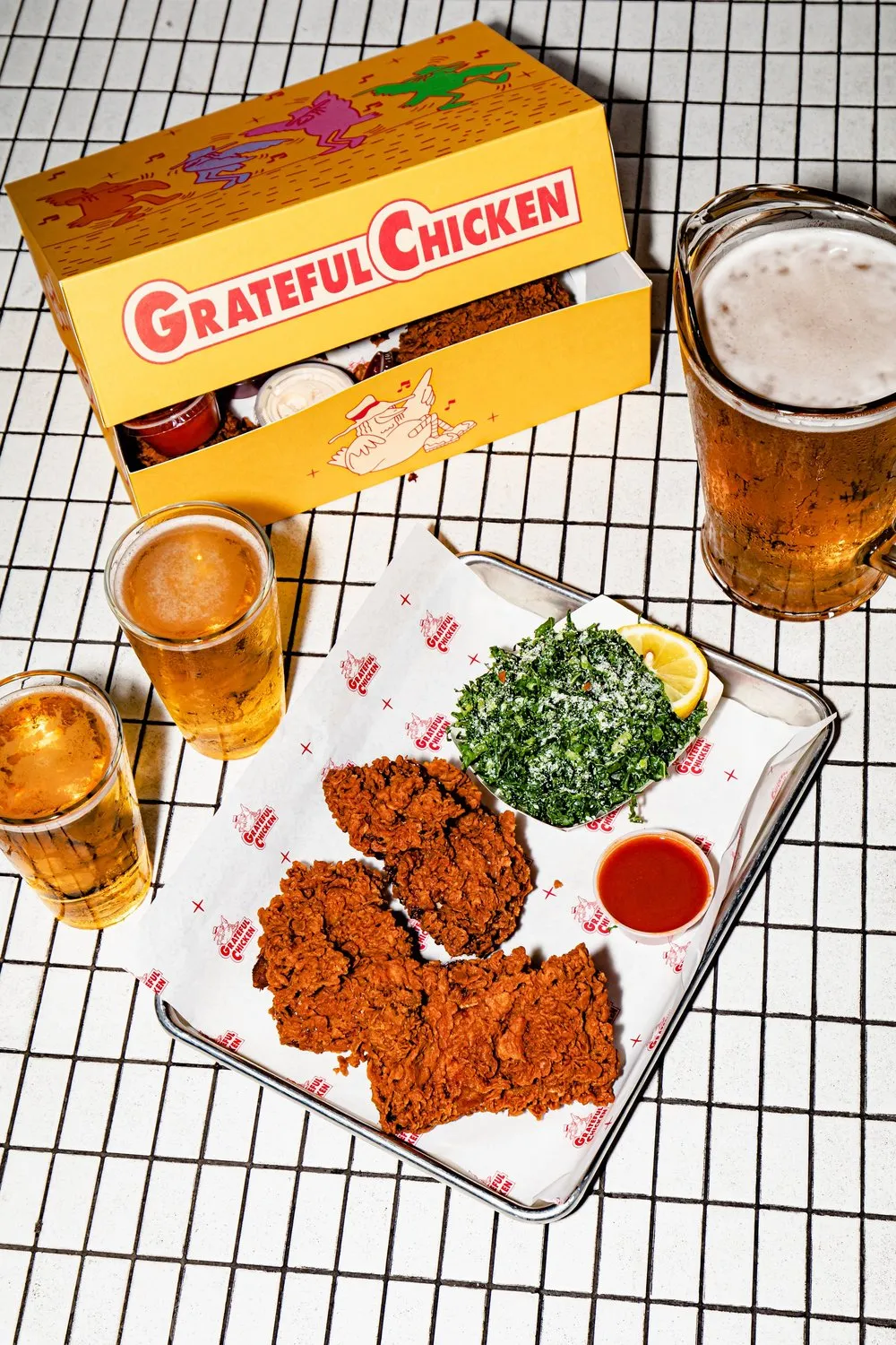 Tray with three pieces of crispy fried chicken, kale salad with lemon wedge, and dipping sauce, accompanied by three glasses of beer and a yellow Grateful Chicken takeout box on a white grid table.