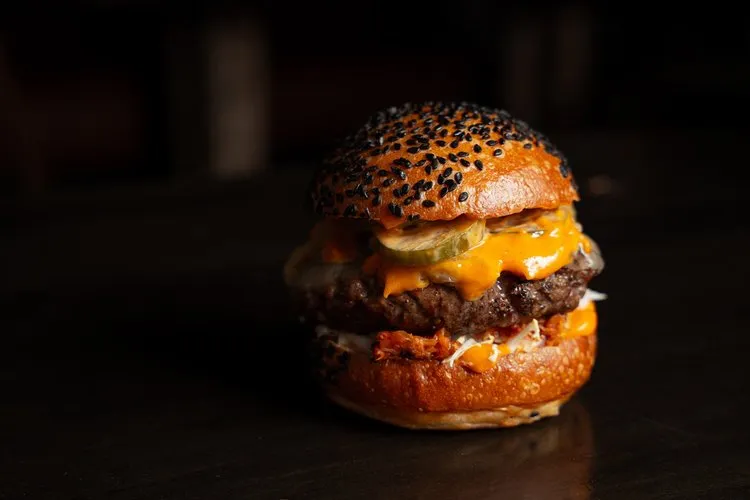 Cheeseburger with melted cheddar, pickles, and bacon on a sesame seed bun against a dark background.