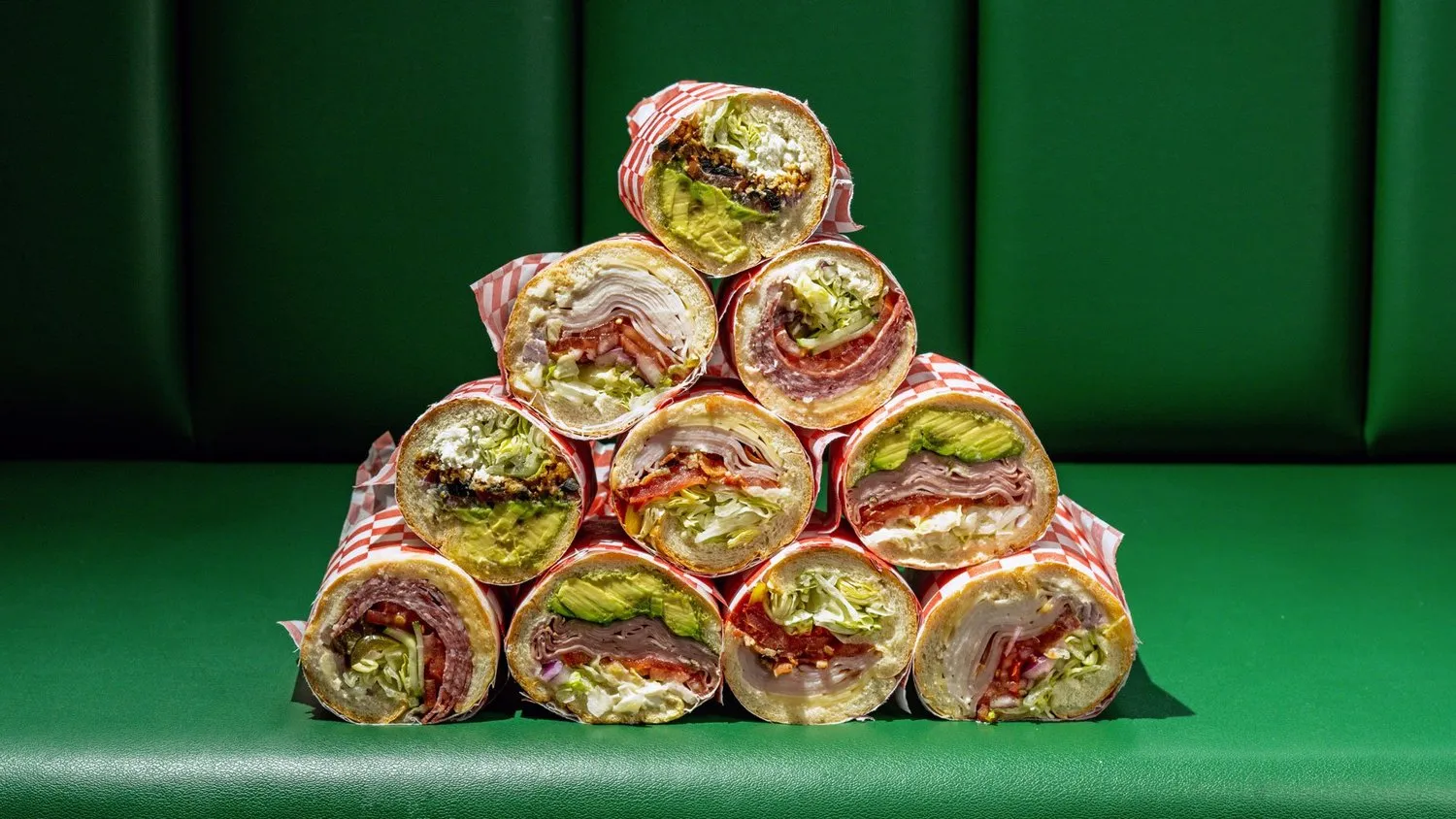 Pyramid stack of nine assorted deli sandwich halves wrapped in red checkered paper on a green leather bench.