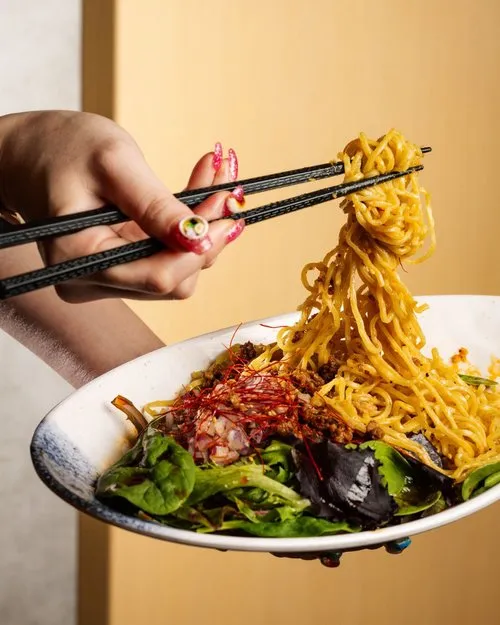 Hand holding black chopsticks lifting yellow ramen noodles from a white bowl with greens and toppings.