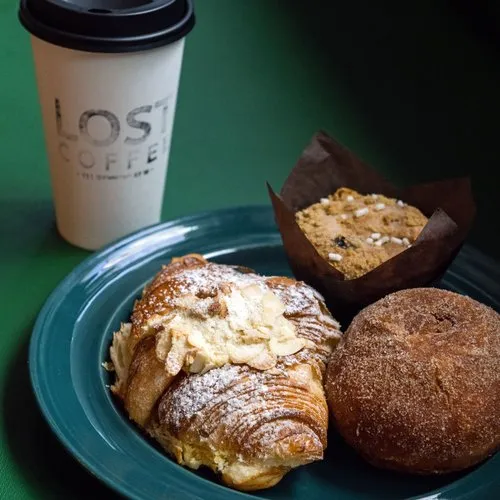 Plate with a powdered sugar croissant filled with cream, a muffin in brown paper, and a cinnamon sugar donut next to a branded coffee cup.