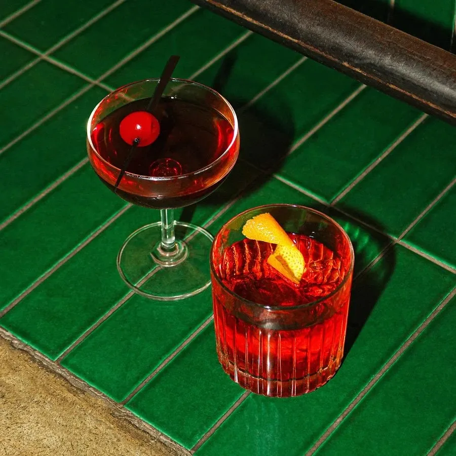 Two red cocktails on green tiled surface, one garnished with a cherry and the other with an orange twist.