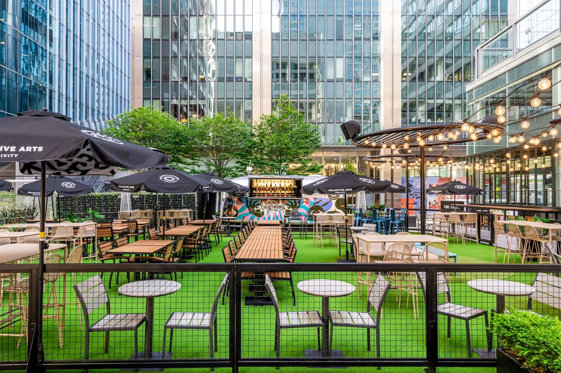 Outdoor urban courtyard patio with tables, chairs, black umbrellas, string lights, and a colorful bar against tall glass buildings.