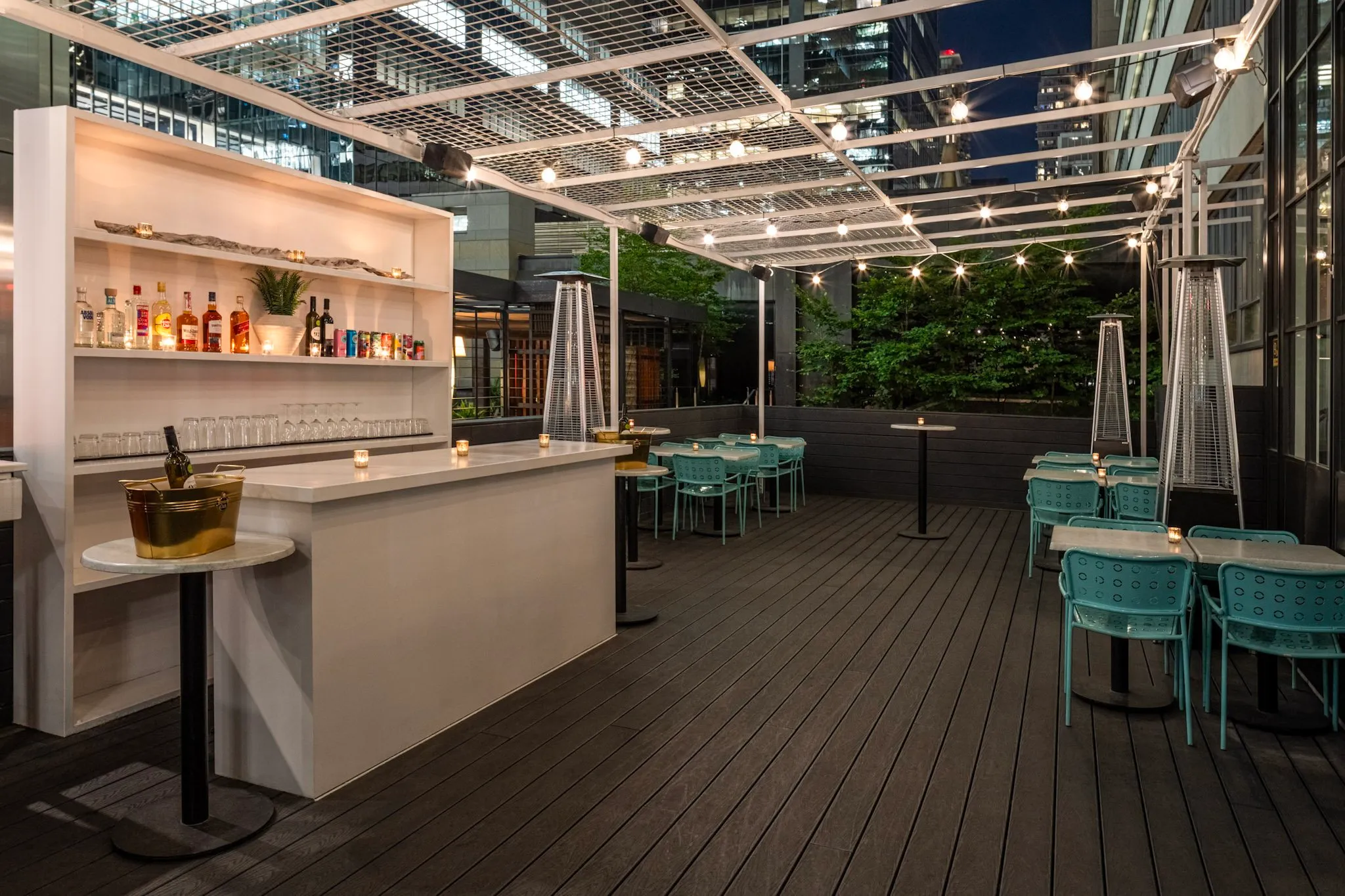 Modern outdoor patio bar at night with string lights, teal chairs, and heaters under a glass canopy.