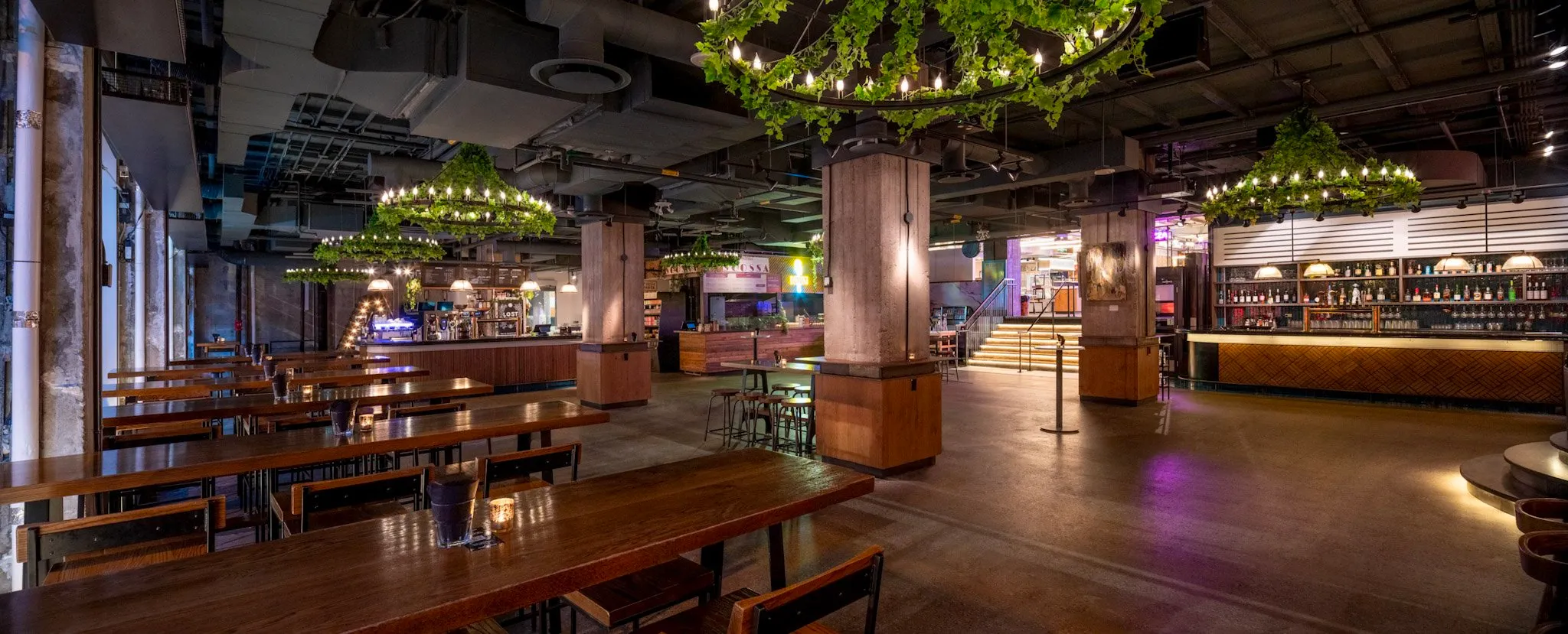 Spacious, modern bar and dining area with long wooden tables, green plant chandeliers, and ambient lighting.