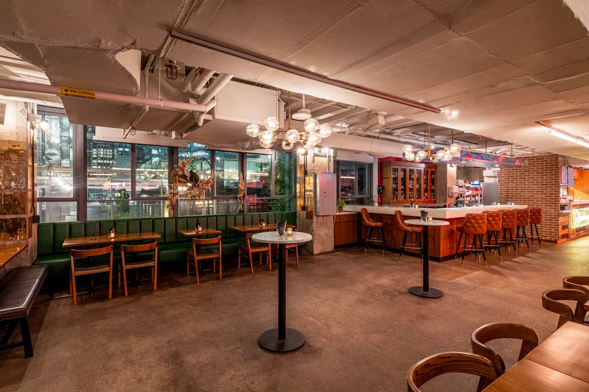 Modern restaurant interior with green banquette seating, wooden chairs, marble-top high tables, and a bar with leather stools under globe chandeliers.