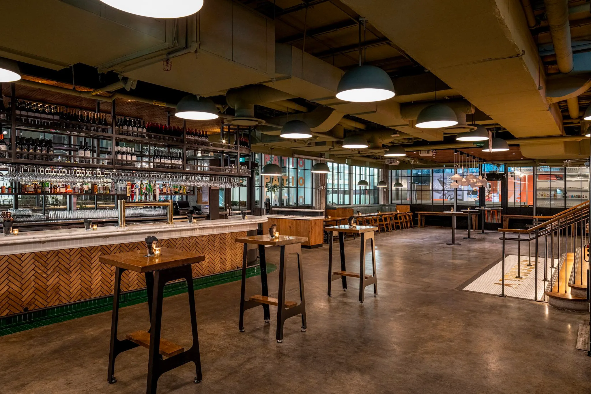Modern empty bar with wooden high tables, a long counter stocked with bottles, hanging wine glasses, and large industrial pendant lights.