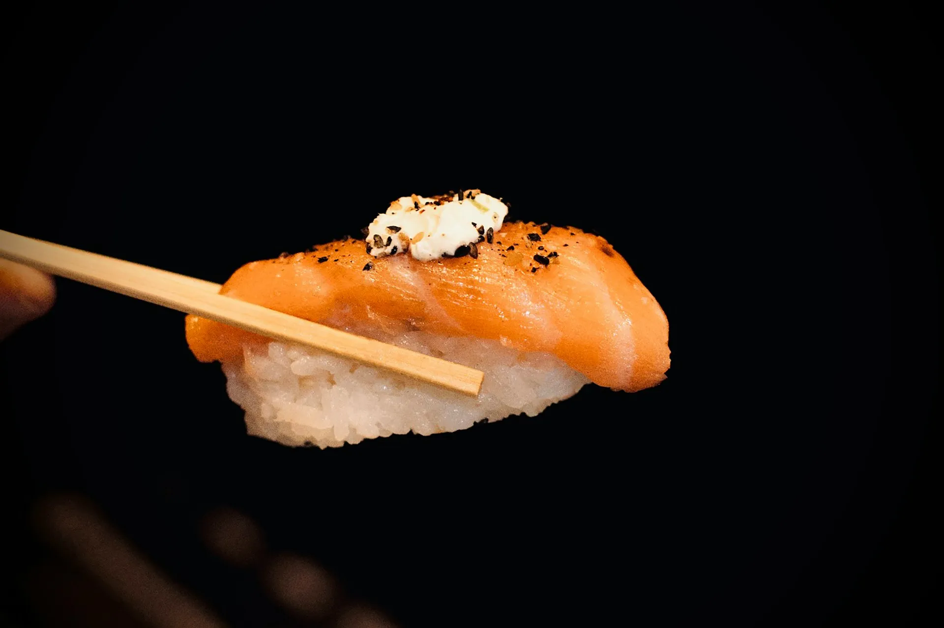 Piece of salmon nigiri sushi topped with cream and black pepper held by chopsticks against a black background.