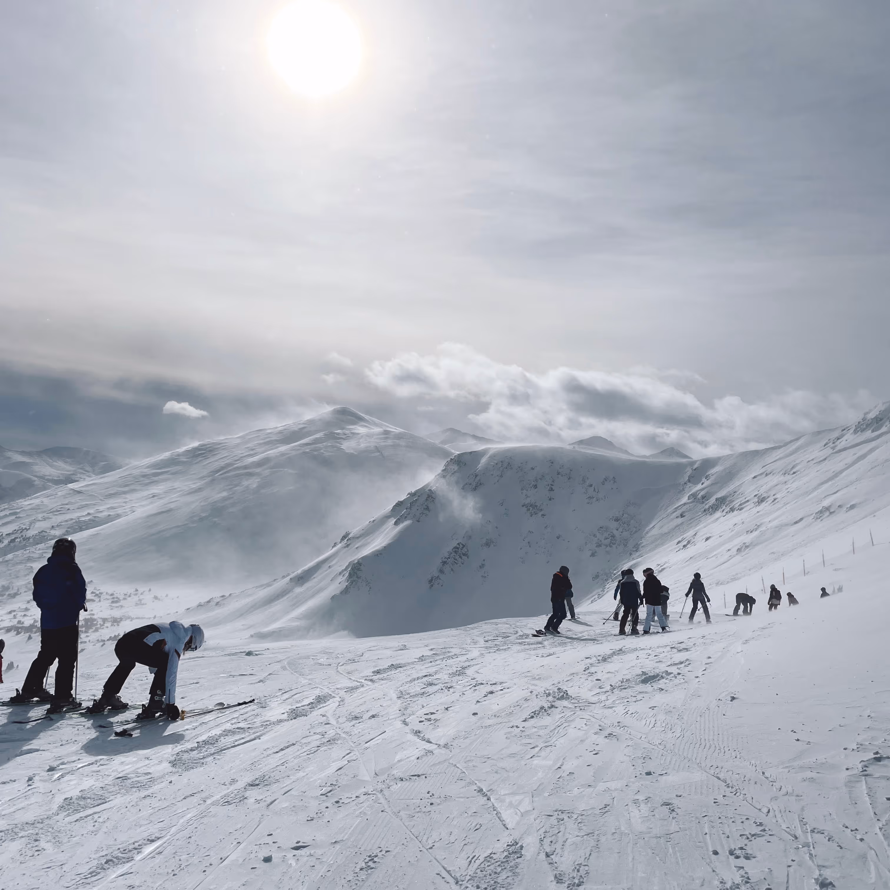 Skiers preparing and skiing on a snowy mountain slope under a bright sun with cloudy sky.