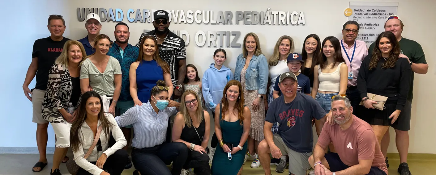 Group of 22 people posing and smiling indoors in front of a sign that reads 'Unidad Cardiovascular Pediátrica Dr. Ortiz'.