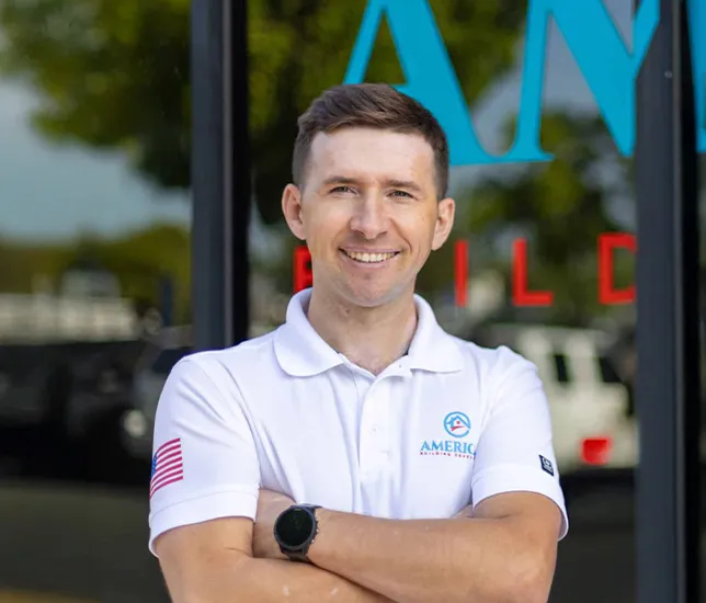 Smiling man with short brown hair wearing a white polo shirt with an American flag patch on the sleeve, standing with arms crossed in front of a glass door.