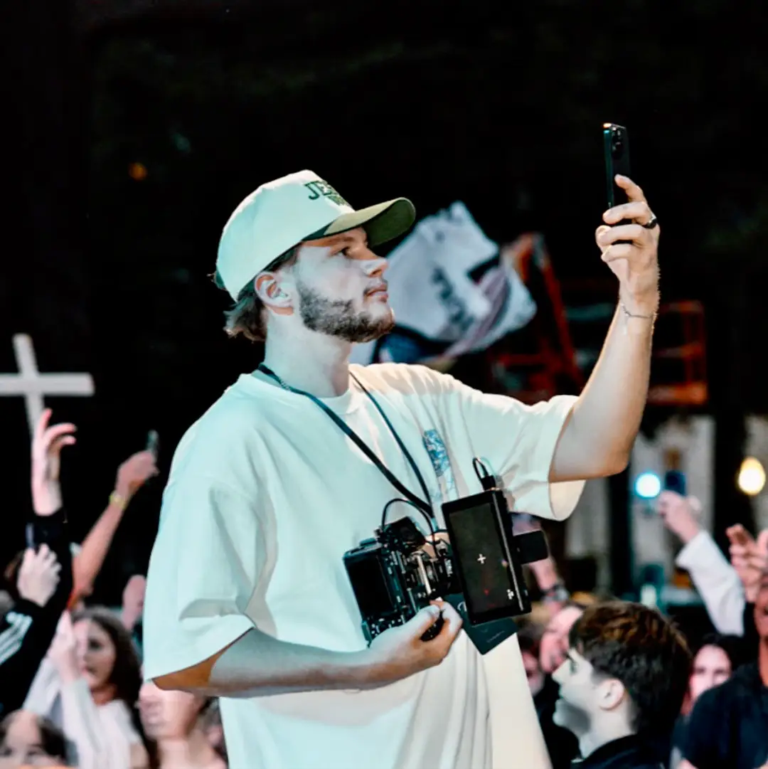 Man wearing a light-colored T-shirt and cap filming with a camera rig while holding a smartphone, with a crowd in the background.