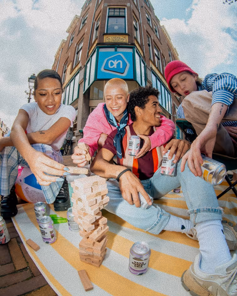 Four friends sitting outside on a blanket playing Jenga and holding canned drinks near a storefront.