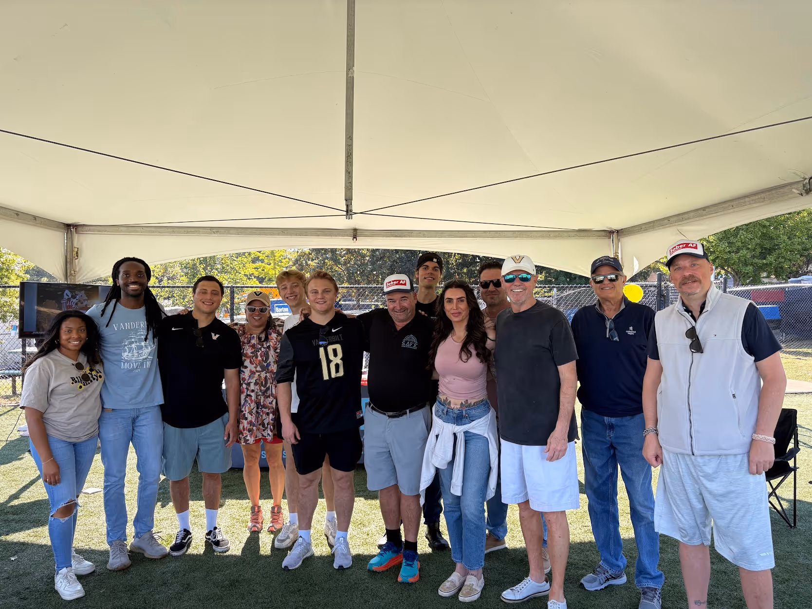 Group of eleven people standing under a white tent on a grassy field, smiling at the camera.