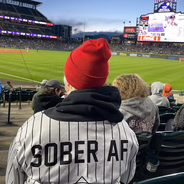 Person wearing a red beanie and a pinstripe jersey with 'SOBER AF' at a baseball stadium.