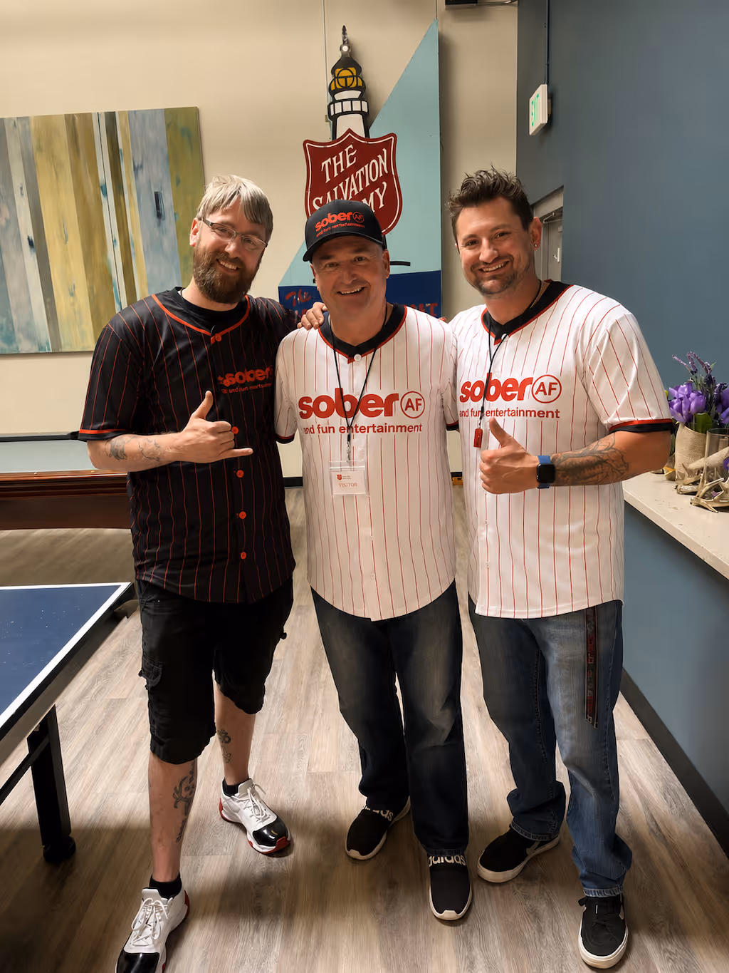 Three men smiling indoors, two in white and one in black 'sober AF' shirts, standing by a Salvation Army sign.