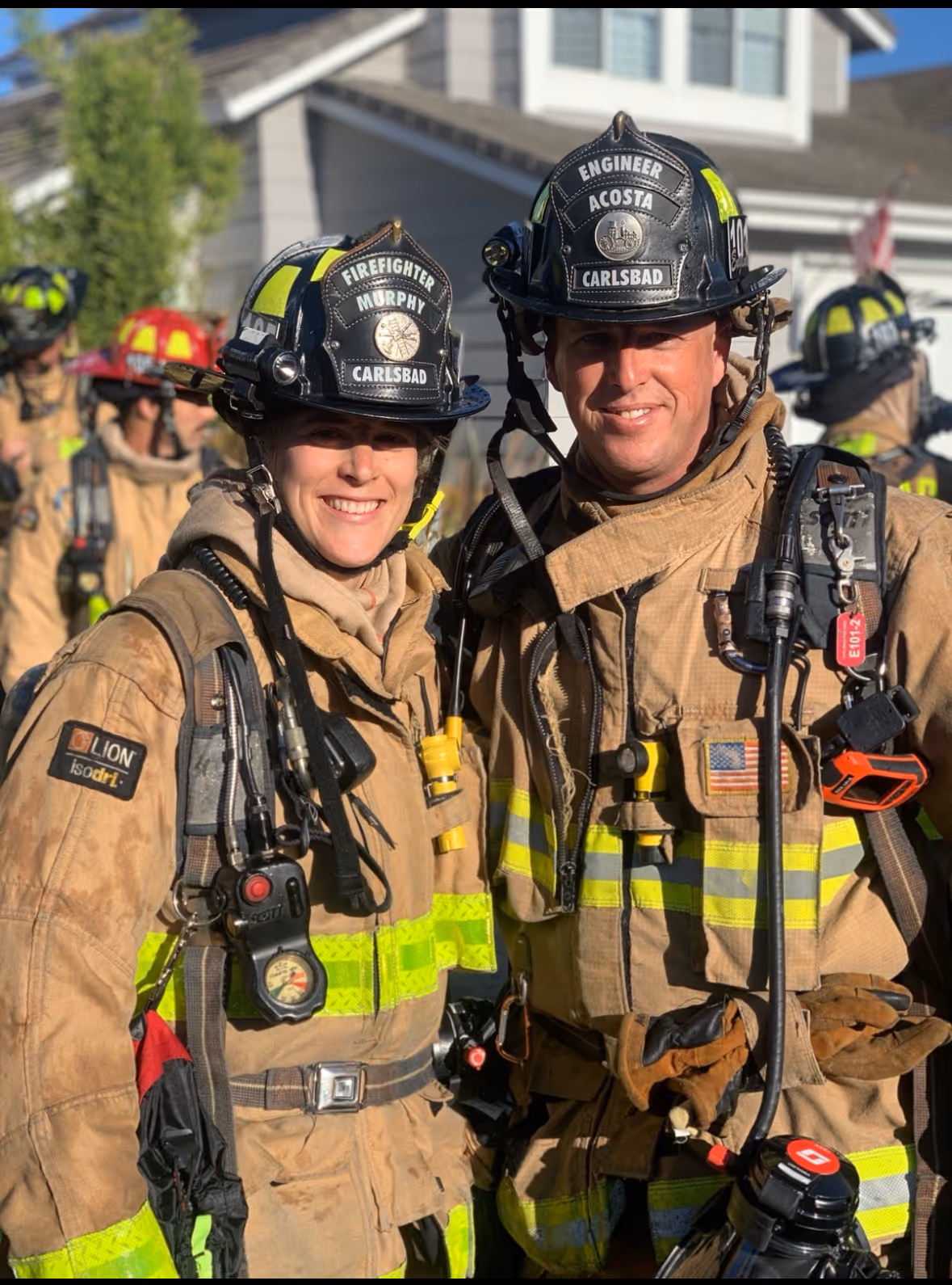 Two firefighters wearing turnout gear and helmets labeled Murphy and Acosta smiling outdoors, with other firefighters in the background.