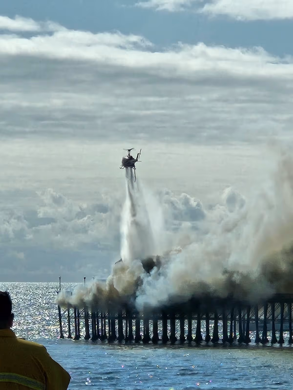 Helicopter spraying water over a large plume of smoke at a pier on a body of water with a person in yellow observing.