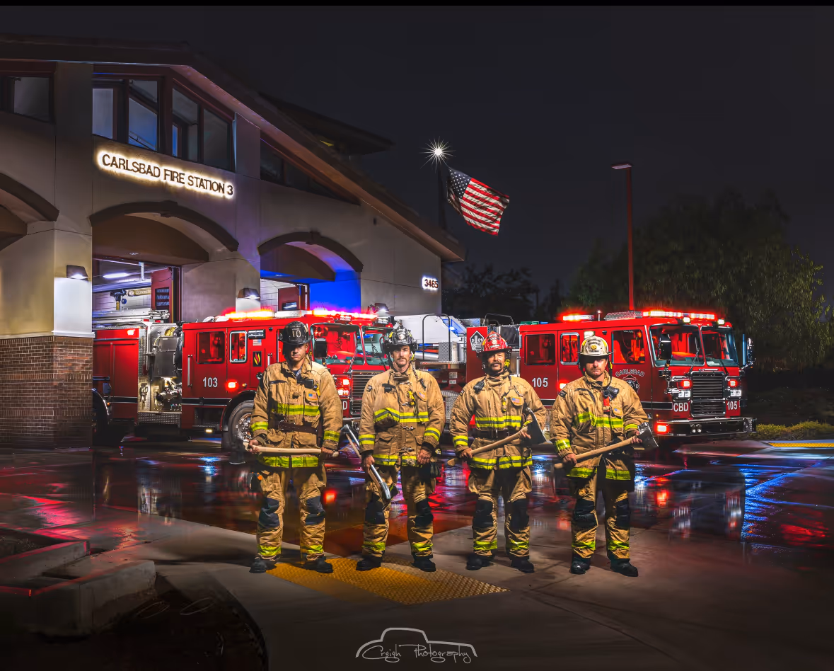 Four firefighters in full gear standing in front of two red fire trucks outside Carlsbad Fire Station 3 at night.