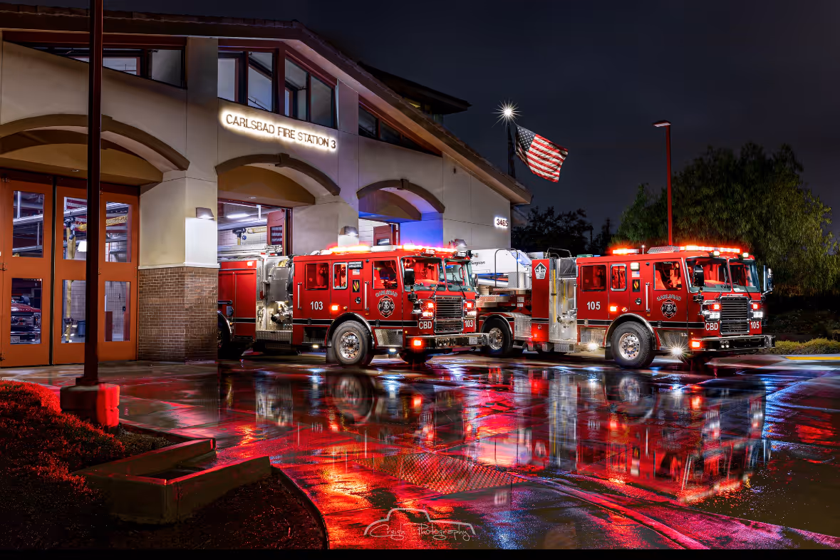 Two red fire trucks parked outside Carlsbad Fire Station 3 at night, reflecting on wet pavement with an American flag waving nearby.