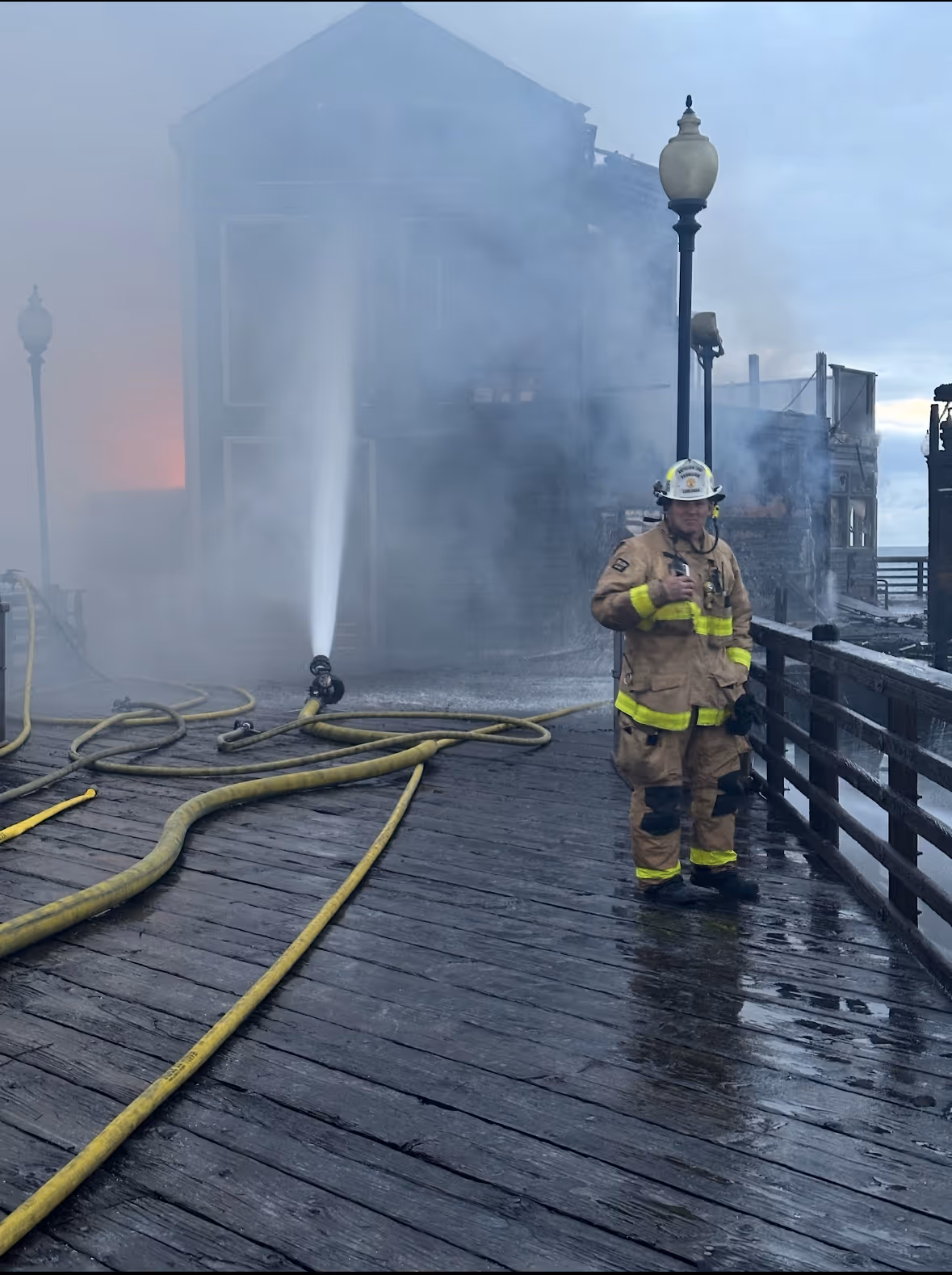 Firefighter in full gear standing on a wet wooden pier with hoses spraying water at a burning building in heavy smoke.