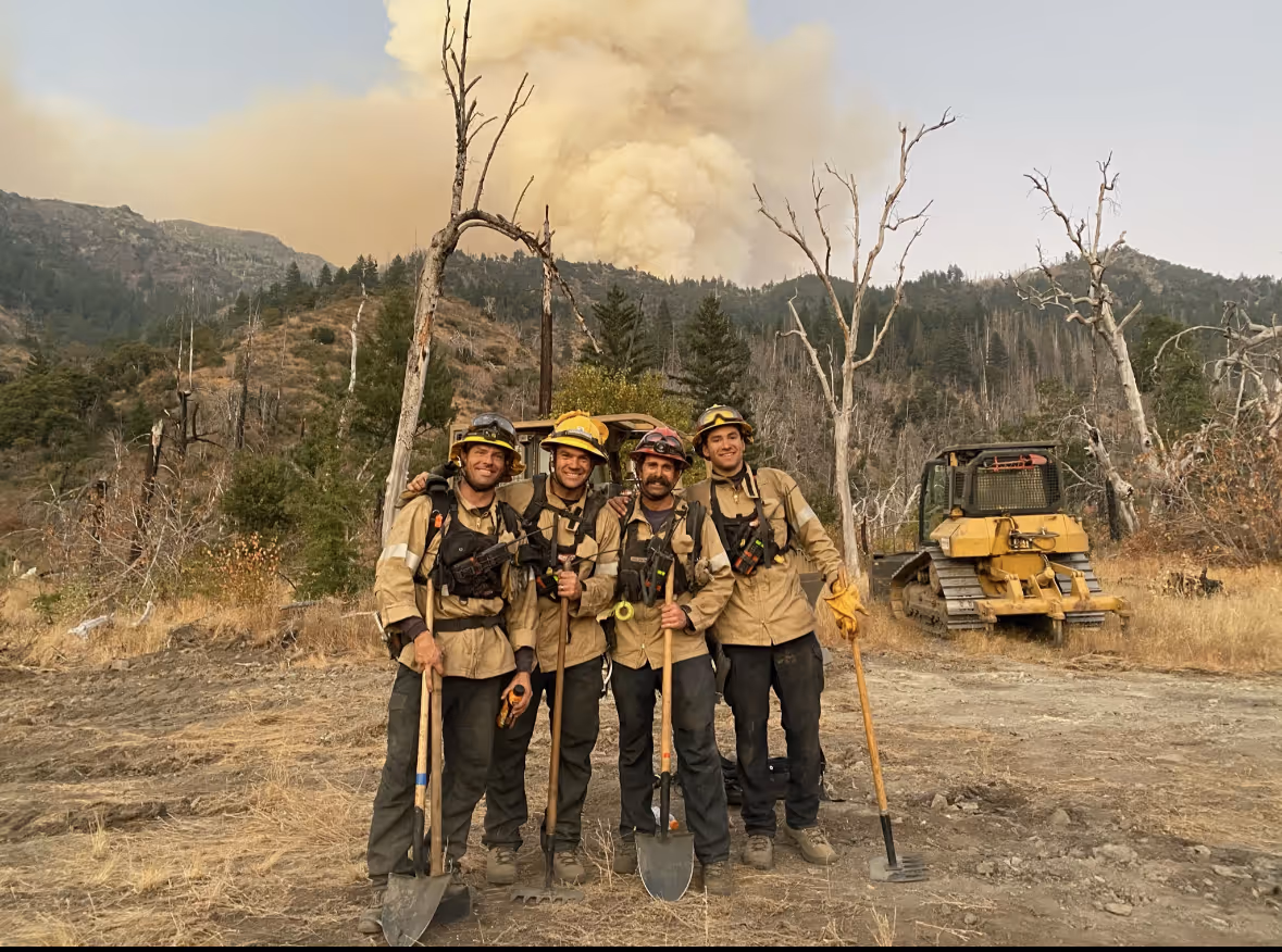 Four firefighters in protective gear posing with tools in a dry forest area with smoke rising from a wildfire behind them.