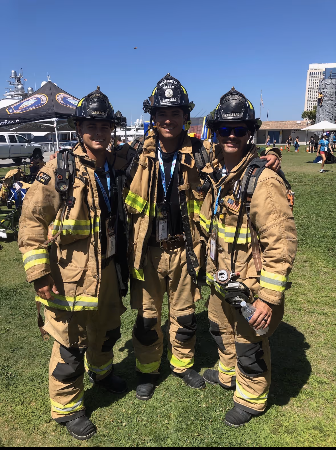 Three firefighters in full gear with helmets and reflective stripes standing arm in arm on grass at an outdoor event on a sunny day.