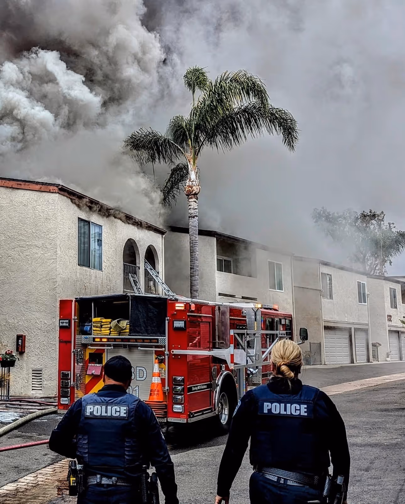 Two police officers walk toward a red fire truck parked near a building emitting thick gray smoke.