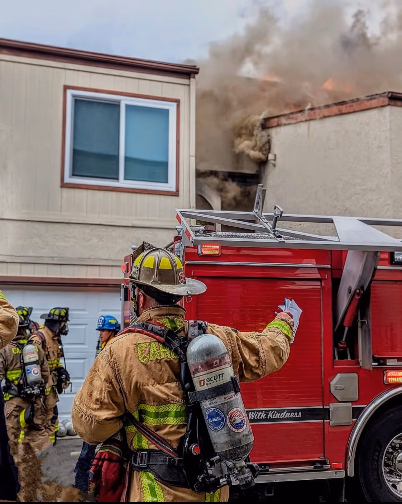 Firefighters in protective gear working near a red fire truck with smoke billowing from a building in the background.