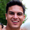 Smiling young man with short dark hair outdoors with greenery in the background.