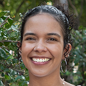 Smiling woman with curly hair wearing a white top standing outdoors with green foliage in the background.