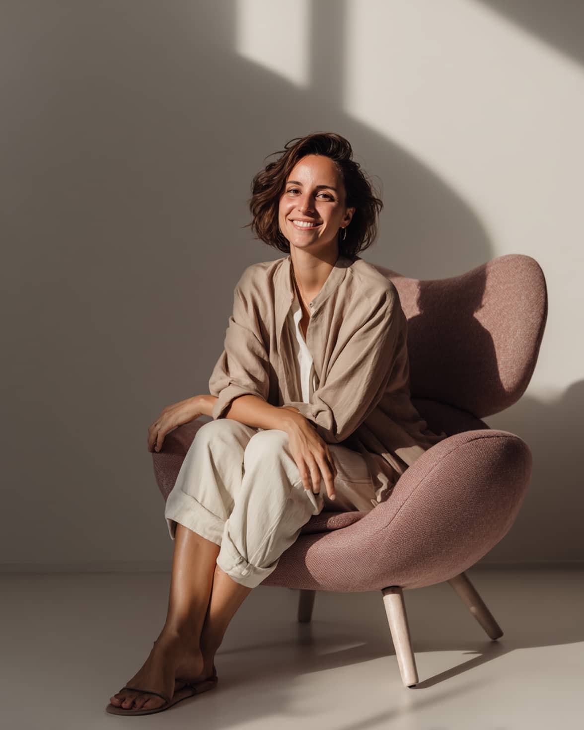 Smiling woman with short brown hair wearing beige and white clothing sitting on a mauve modern armchair in soft natural light.