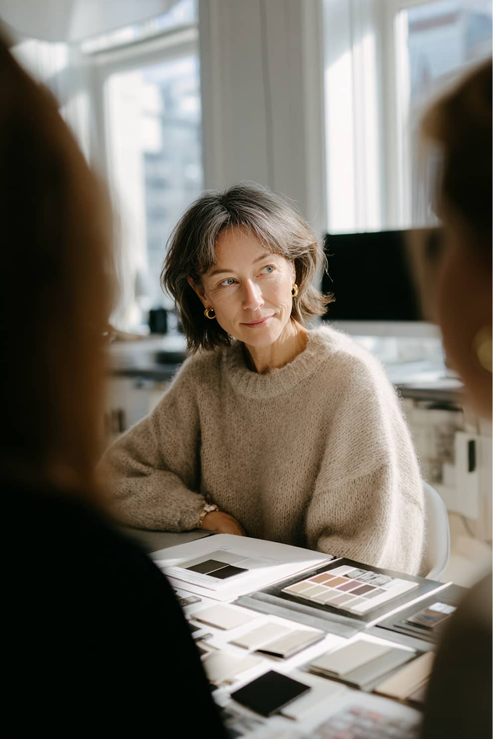 Middle-aged woman in a beige sweater sitting at a table with color and fabric samples, engaged in discussion.