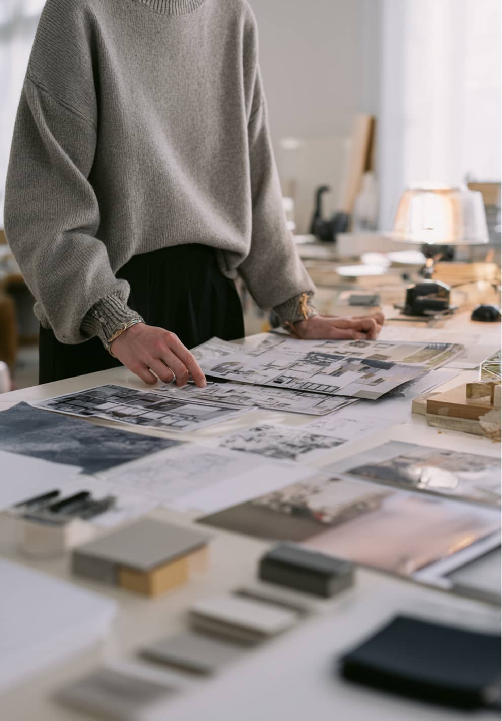 Person in a gray sweater reviewing architectural blueprints and design plans on a desk covered with samples and sketches.