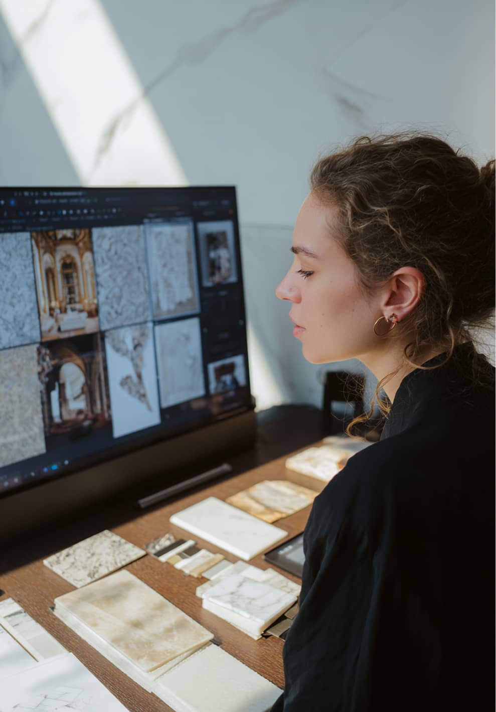 Woman with curly hair observing marble and tile samples arranged on a desk and displayed on a large monitor.