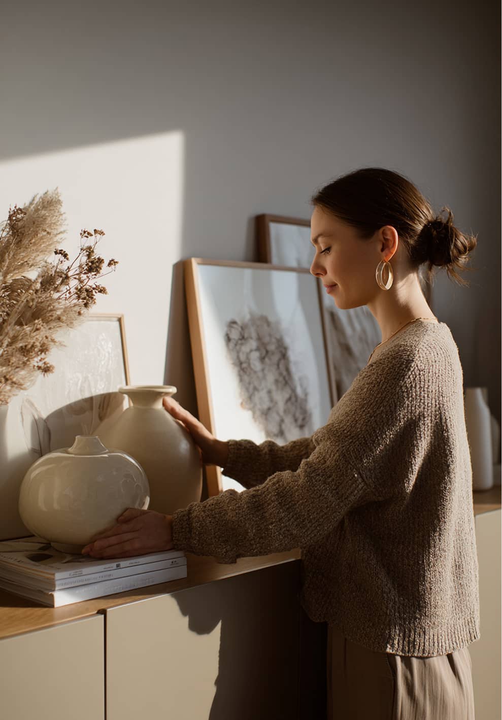 A woman arranging beige ceramic vases on a wooden cabinet with framed artwork and dried flowers behind her.