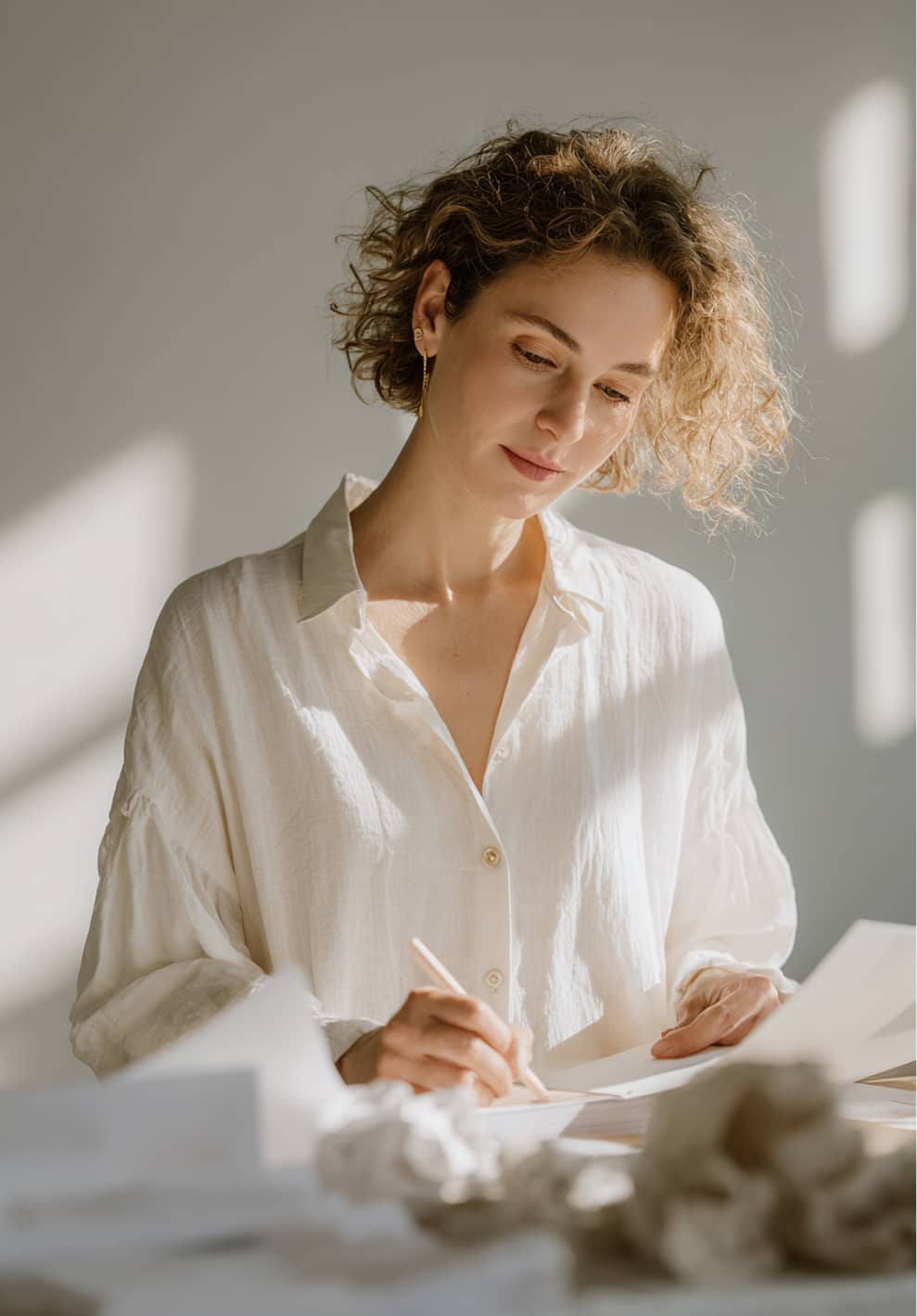 Woman with curly hair in a white shirt writing on paper at a sunlit desk.