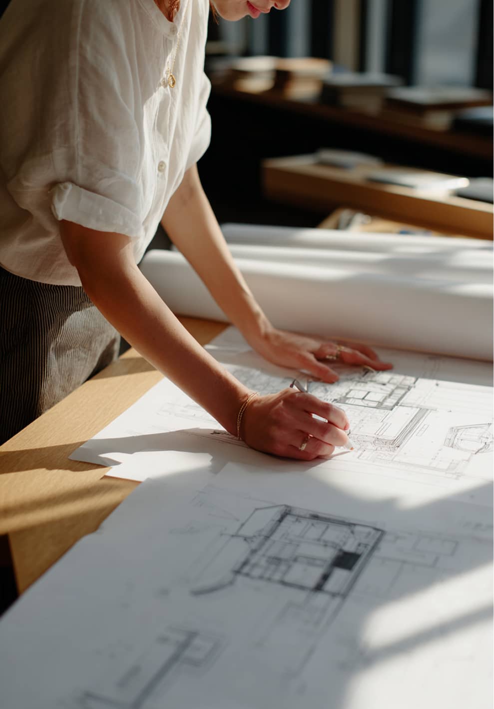 Person sketching architectural blueprints on a wooden table in natural light.