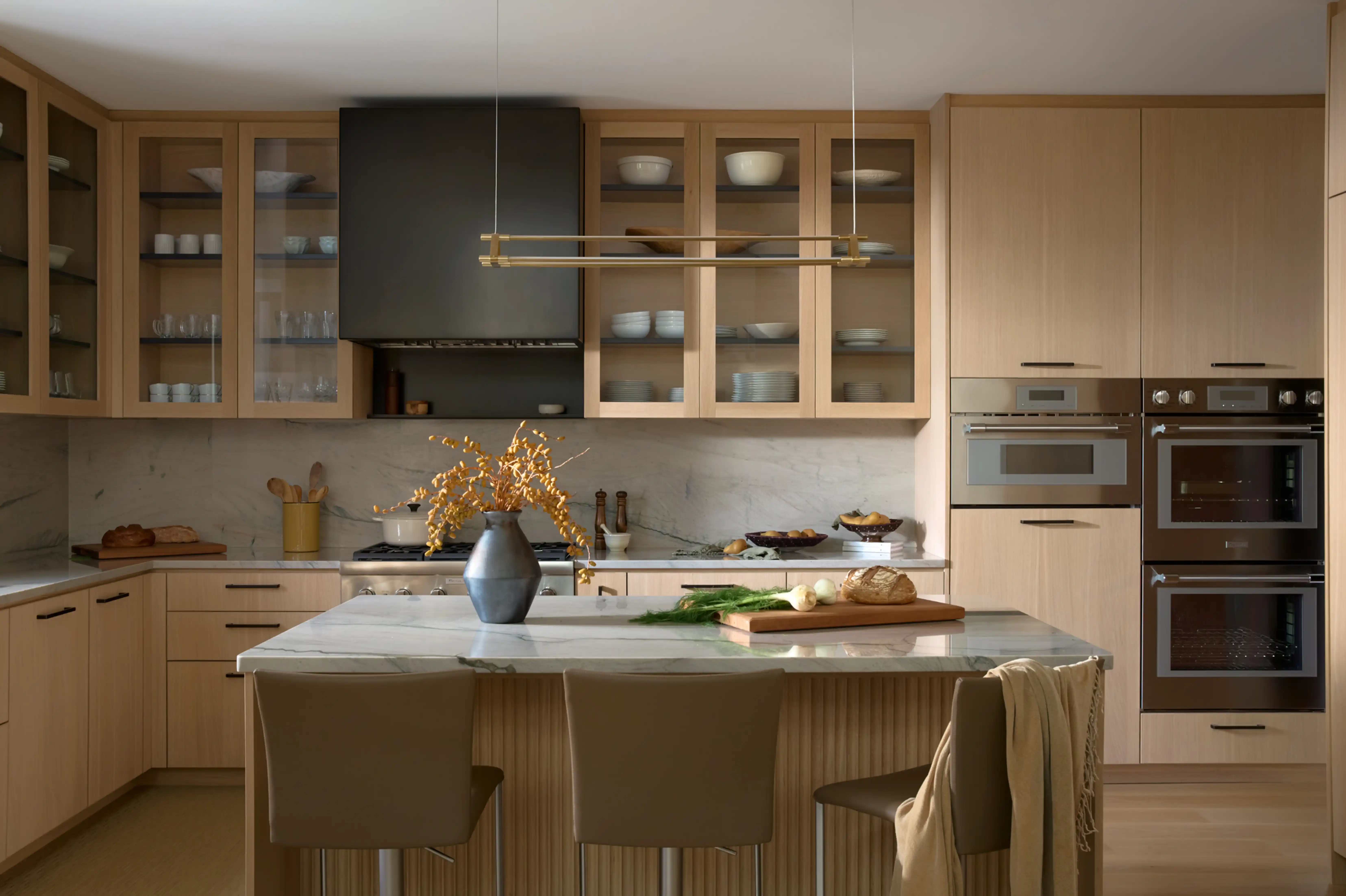 Modern kitchen with light wood cabinets, marble countertops, glass-fronted upper cabinets, and an island with three chairs and decorative vase.