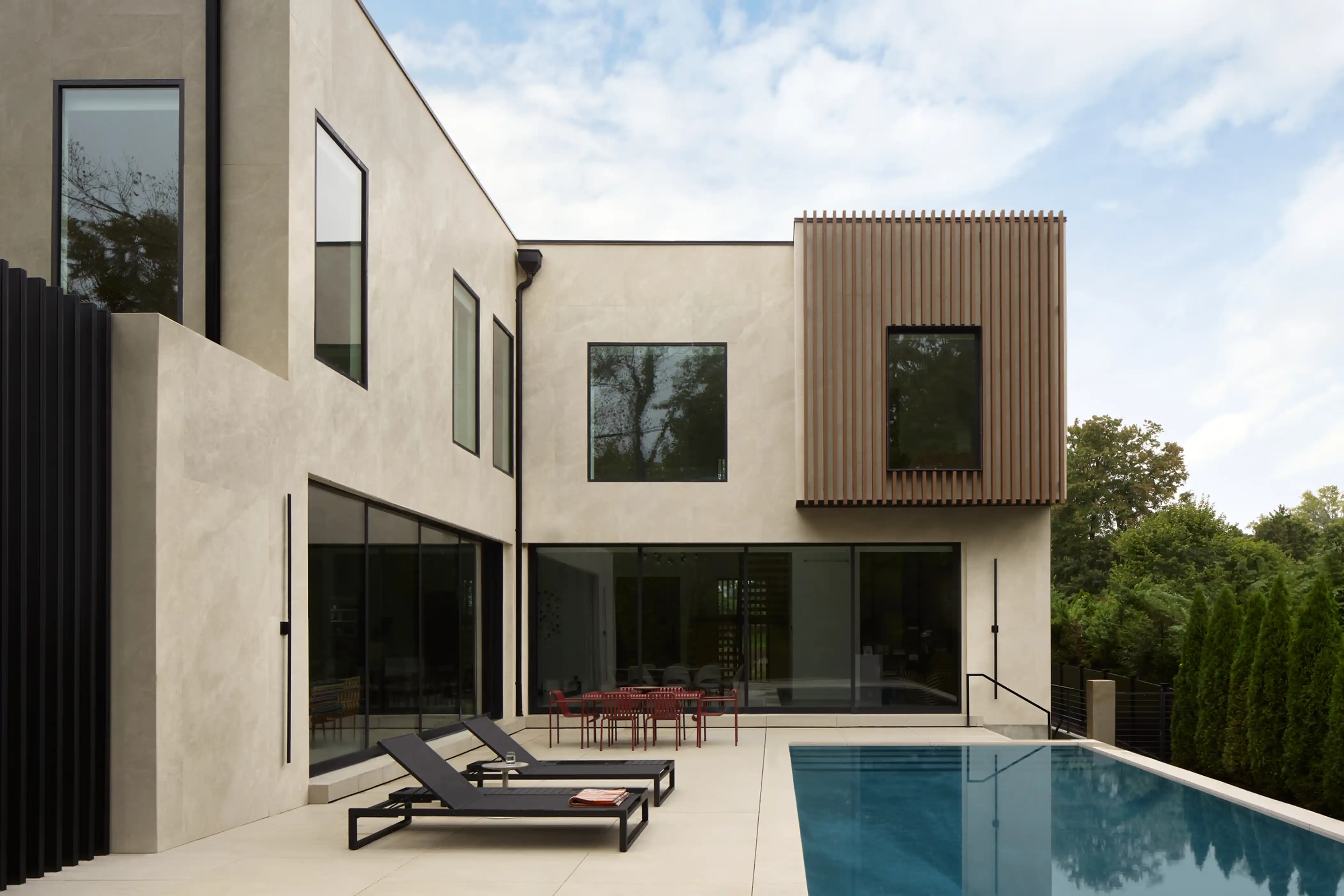 Modern two-story house with beige stucco exterior, large windows, an outdoor pool, two black lounge chairs, and a red dining set on the patio.