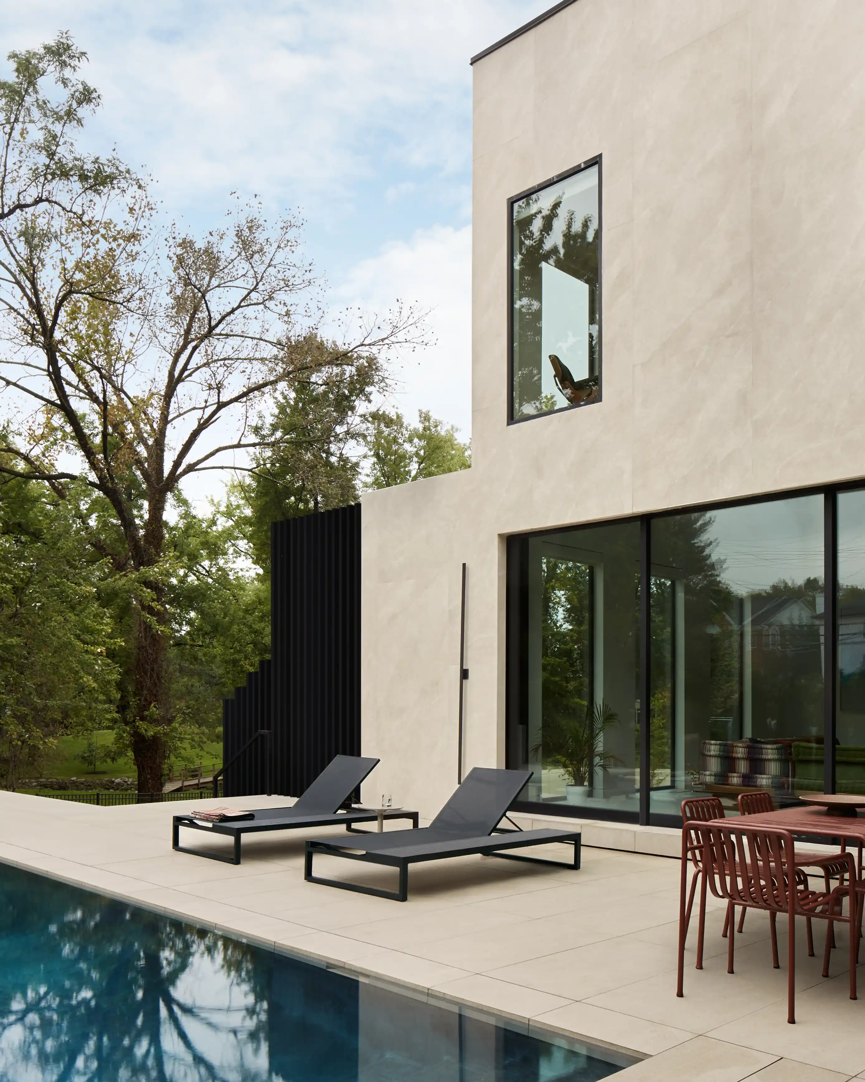 Modern backyard with two dark gray lounge chairs beside a swimming pool and a patio table with chairs next to a beige house with large windows.
