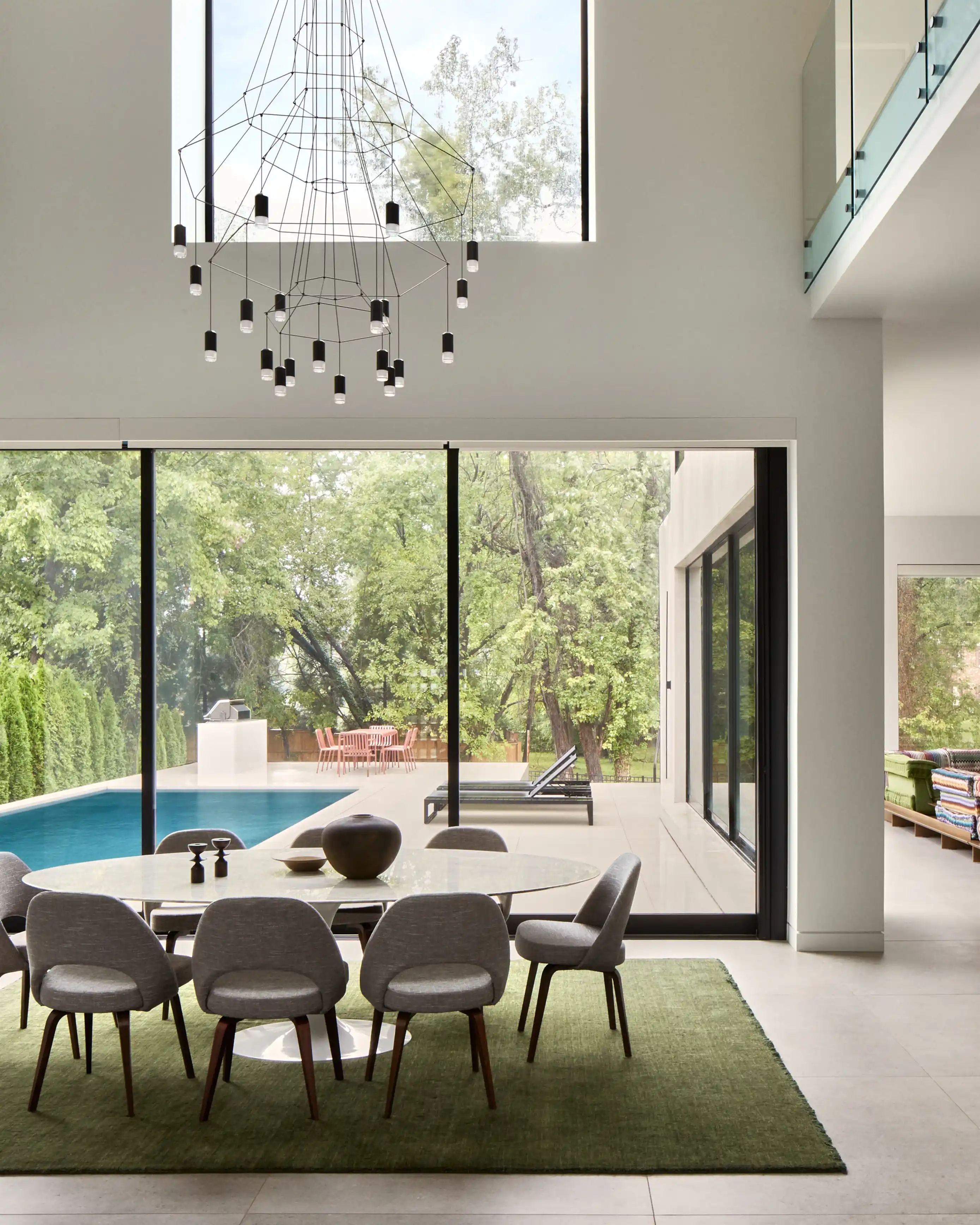 Modern dining area with oval table, eight gray chairs on a green rug, large floor-to-ceiling windows overlooking a pool and outdoor patio with trees.