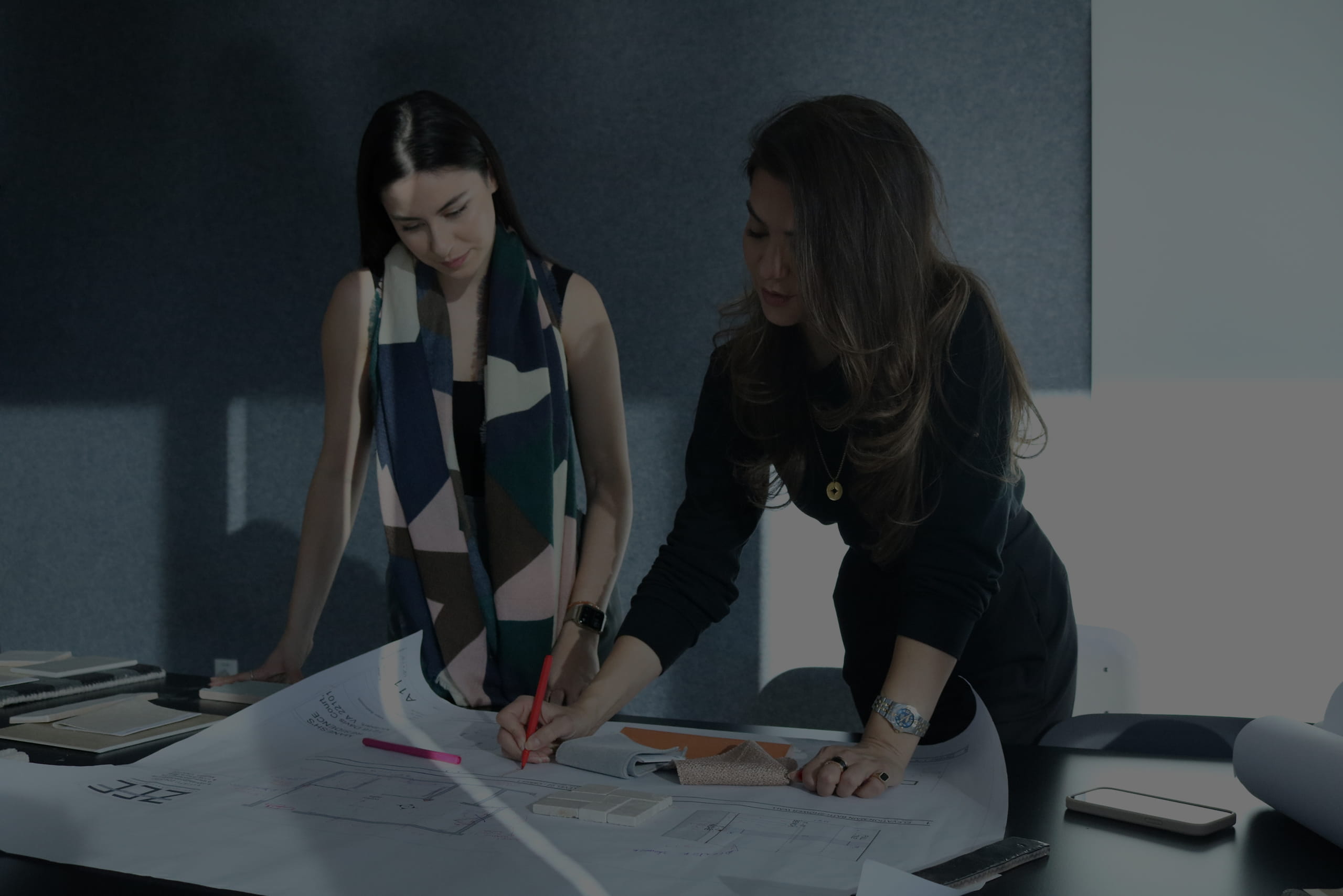 Two women collaborating over architectural plans on a table, one writing with a red pen, the other observing.