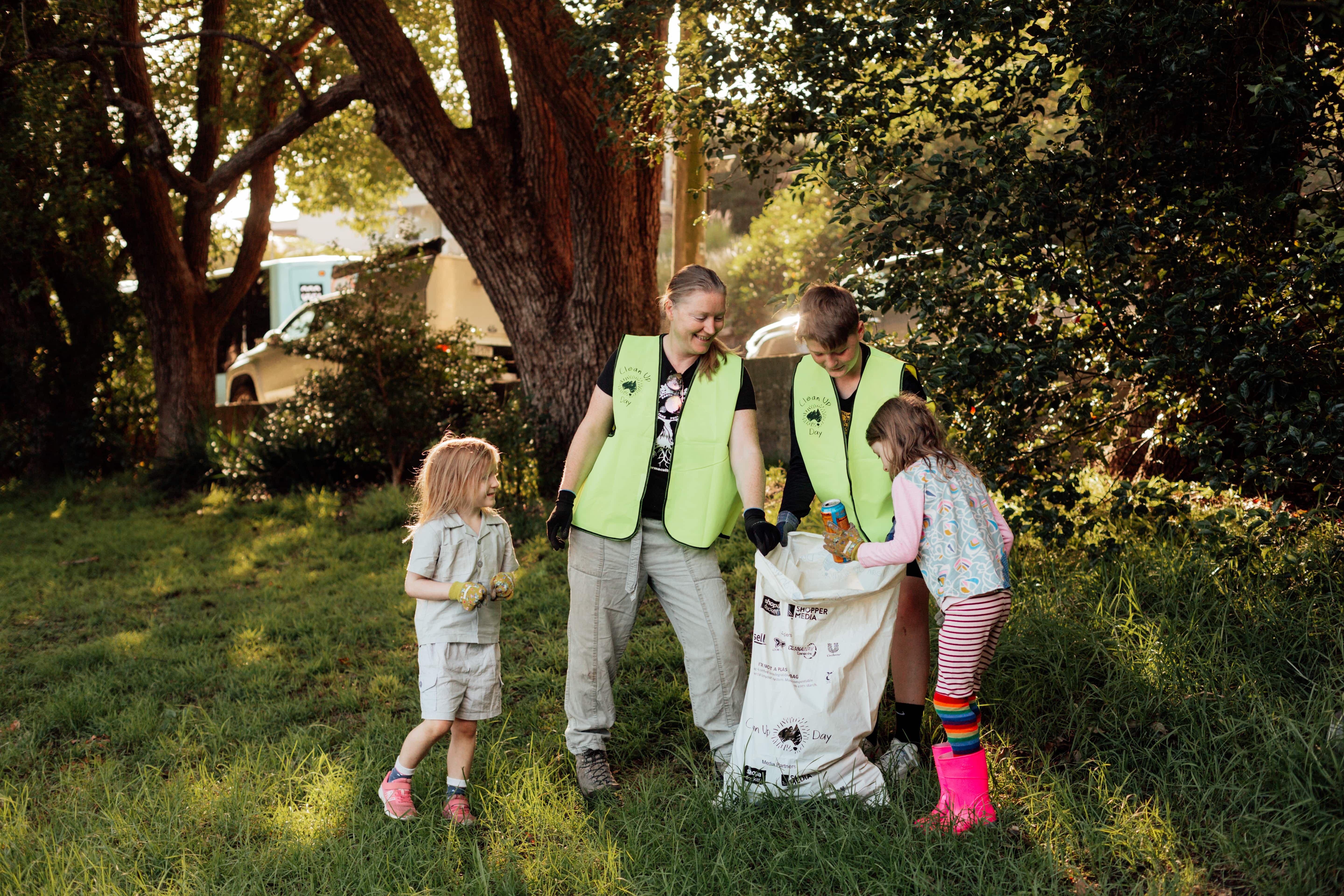 An adult and three children in a park. They are wearing gloves and putting litter in a large rubbish bag.