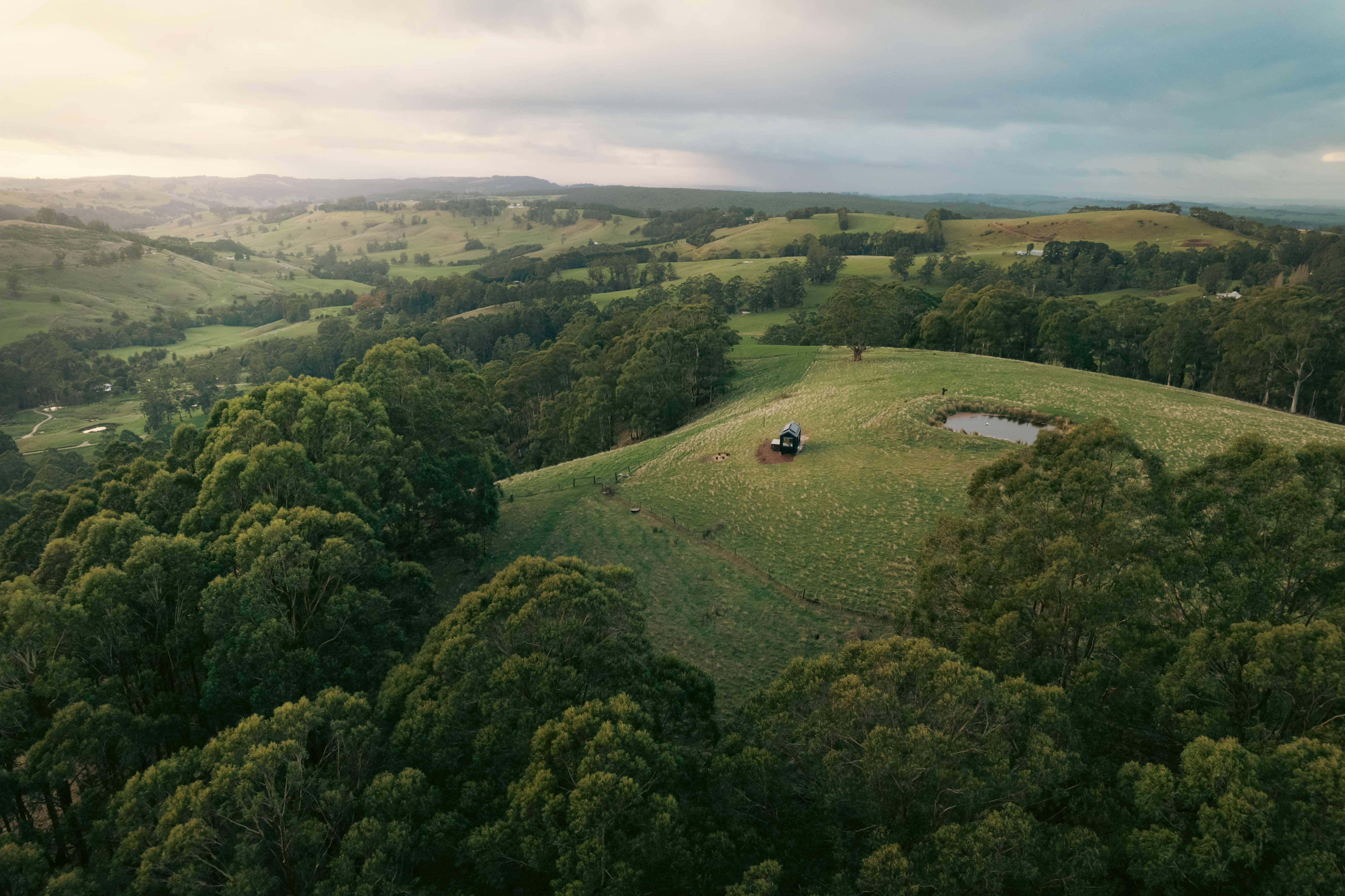 A small cottage in a green paddock on top of a hill surrounded by bushland.