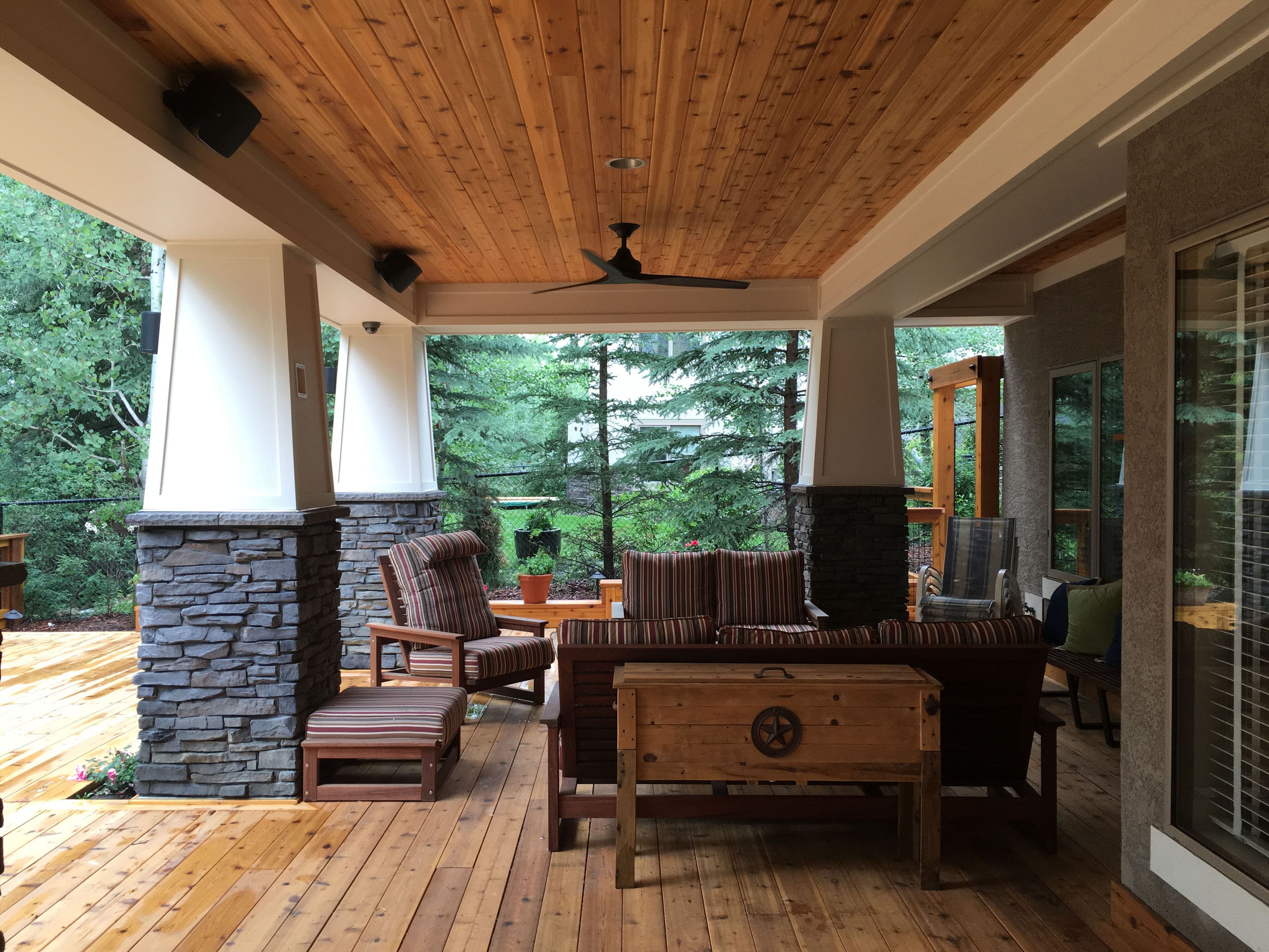 Covered wooden porch with stone pillars, striped cushioned furniture, ceiling fan, and view of green trees.
